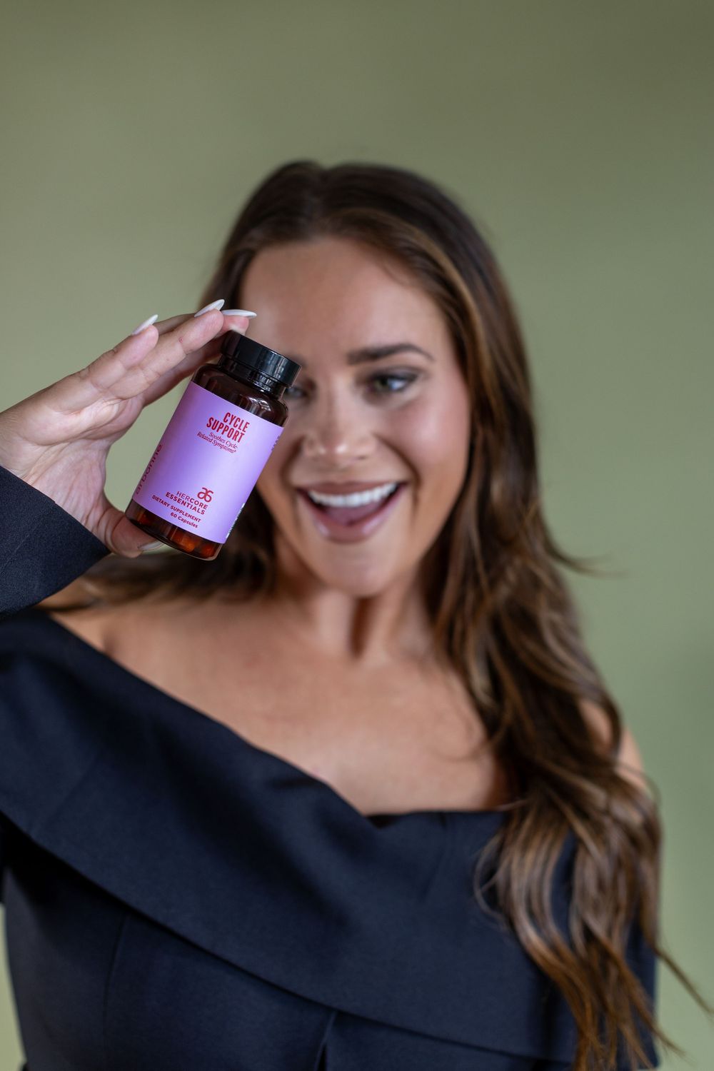 Woman in black off-shoulder top holds up purple vitamin supplement bottle while smiling against green background.