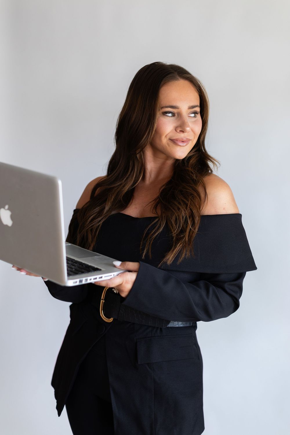Young professional in black off-shoulder outfit smiling while holding MacBook laptop against light gray background.