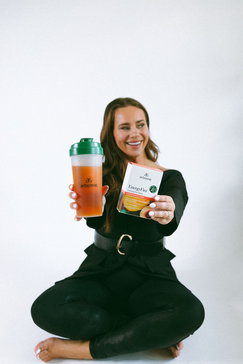 Person in black clothing holds up orange and green products while sitting cross-legged on white background.
