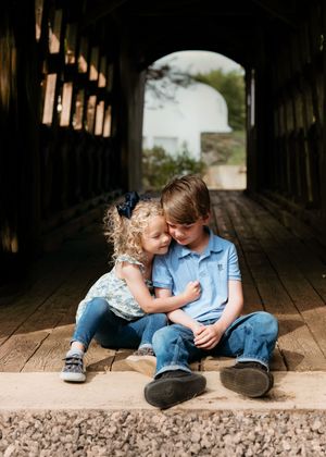 Two young siblings sitting on a wooden covered bridge floor, with the girl hugging her older brother.
