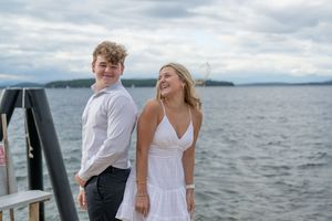 A young couple smiles on a dock by a lake, wearing white outfits on a cloudy day.