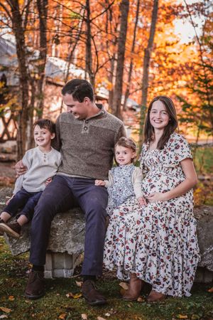A smiling pregnant woman sits with her family outdoors amid vibrant autumn foliage.