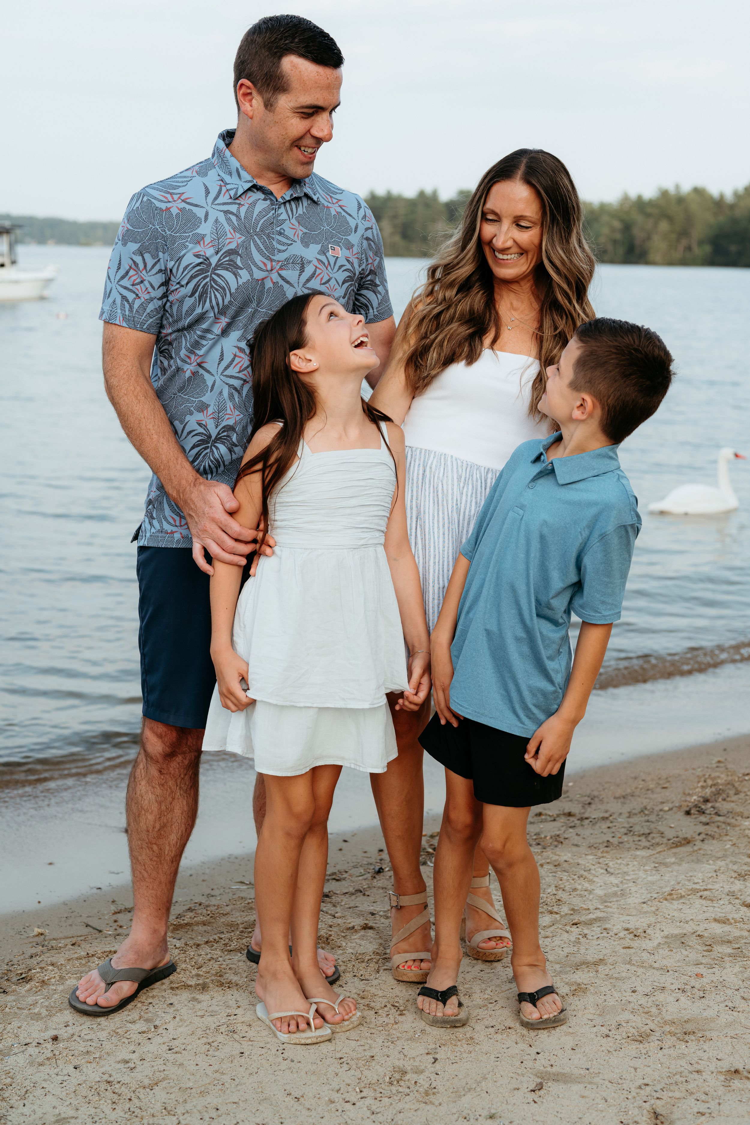 A happy family of four poses on a sandy beach by a calm lake at sunset.