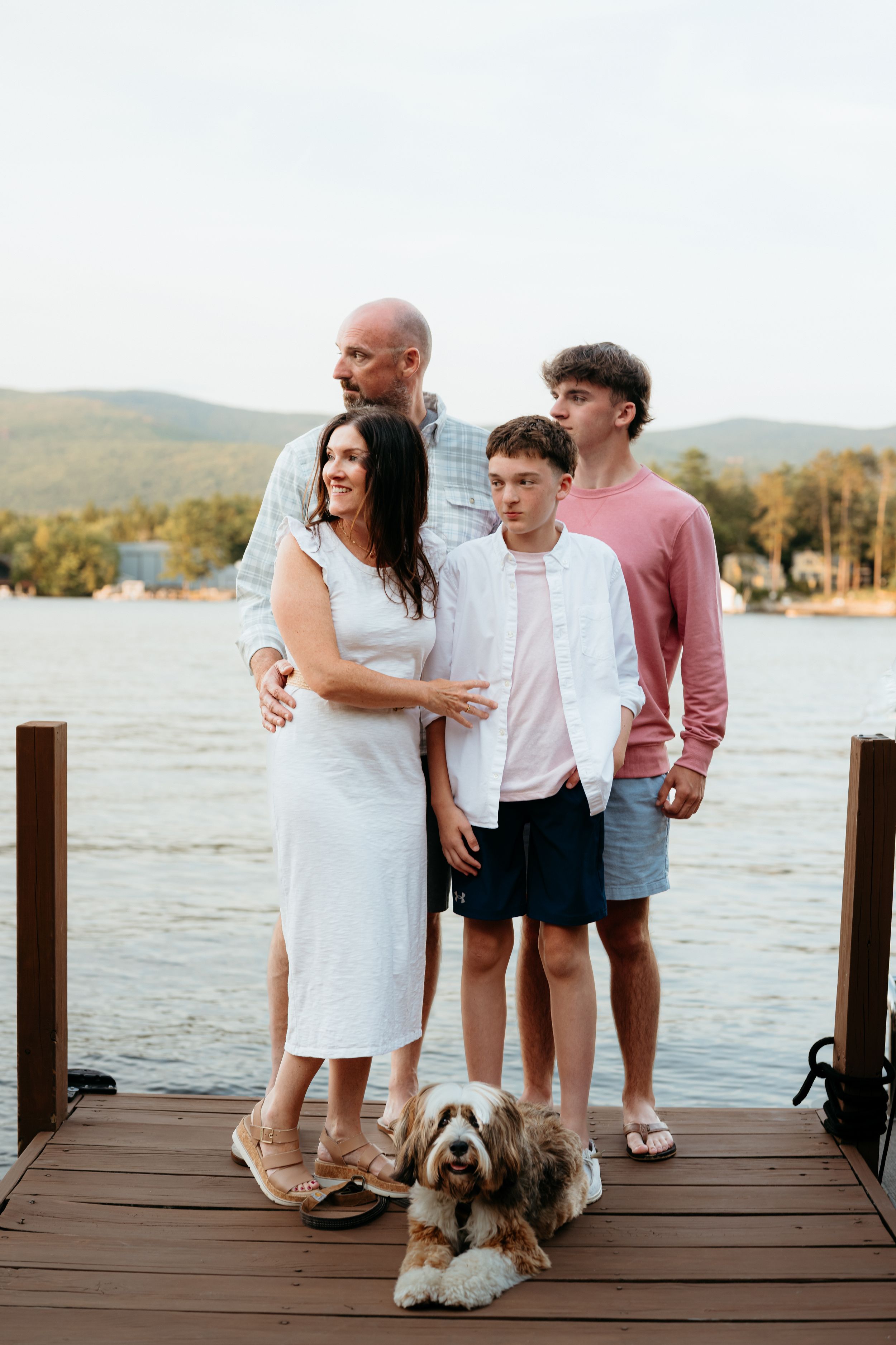 Family of four with a fluffy dog posing on a lakeside dock with mountains in the background.
