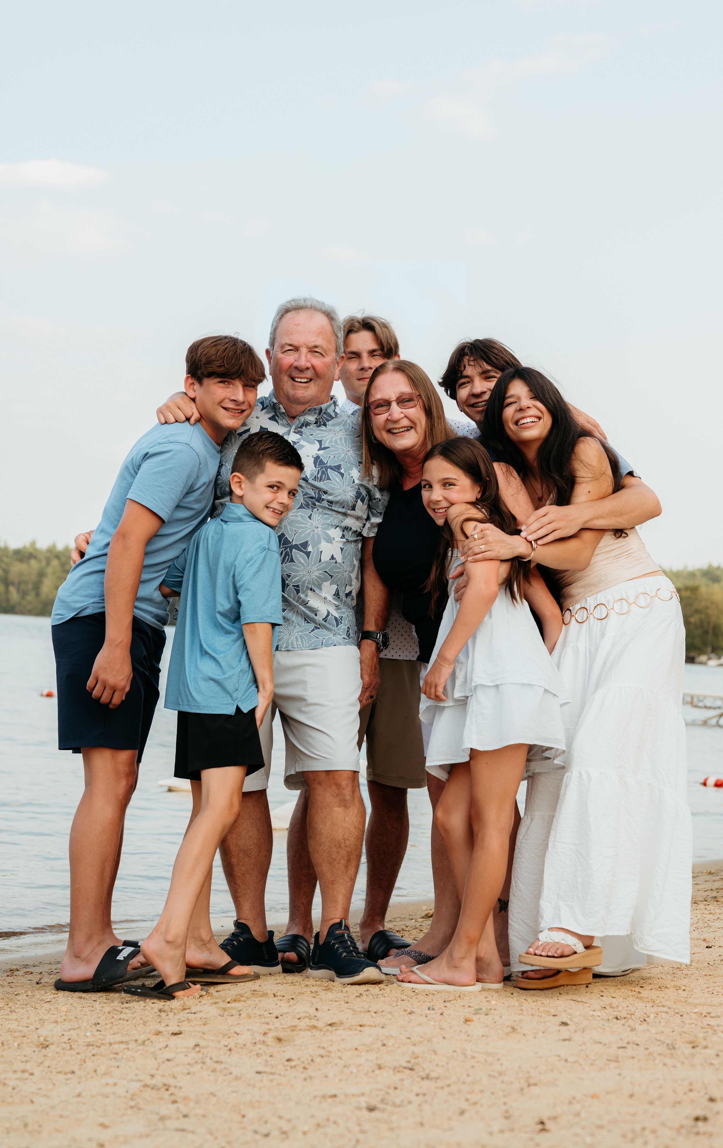 A large multigenerational family group hugs and smiles together on a sunny beach.