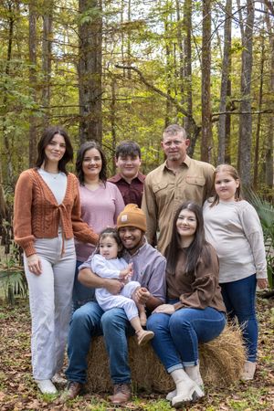 A family poses together on hay bales in a sunlit autumn forest wearing coordinated casual outfits.