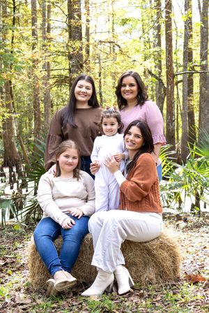 Family group sits casually arranged on hay bales in a wooded setting with golden sunlight filtering through trees.