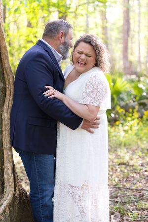 Couple embracing and laughing in a sunlit forest, woman in white lace dress, man in navy blazer and jeans.