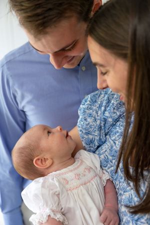 Young parents in blue attire lovingly gaze at their newborn baby dressed in a white smocked outfit.