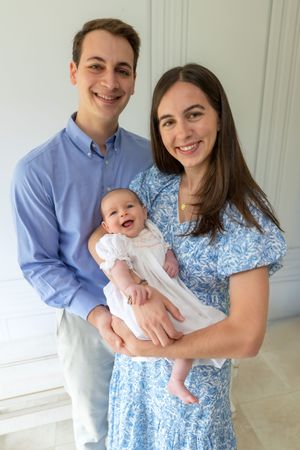 Happy young family portrait with smiling parents in blue outfits holding their laughing newborn baby in white dress.