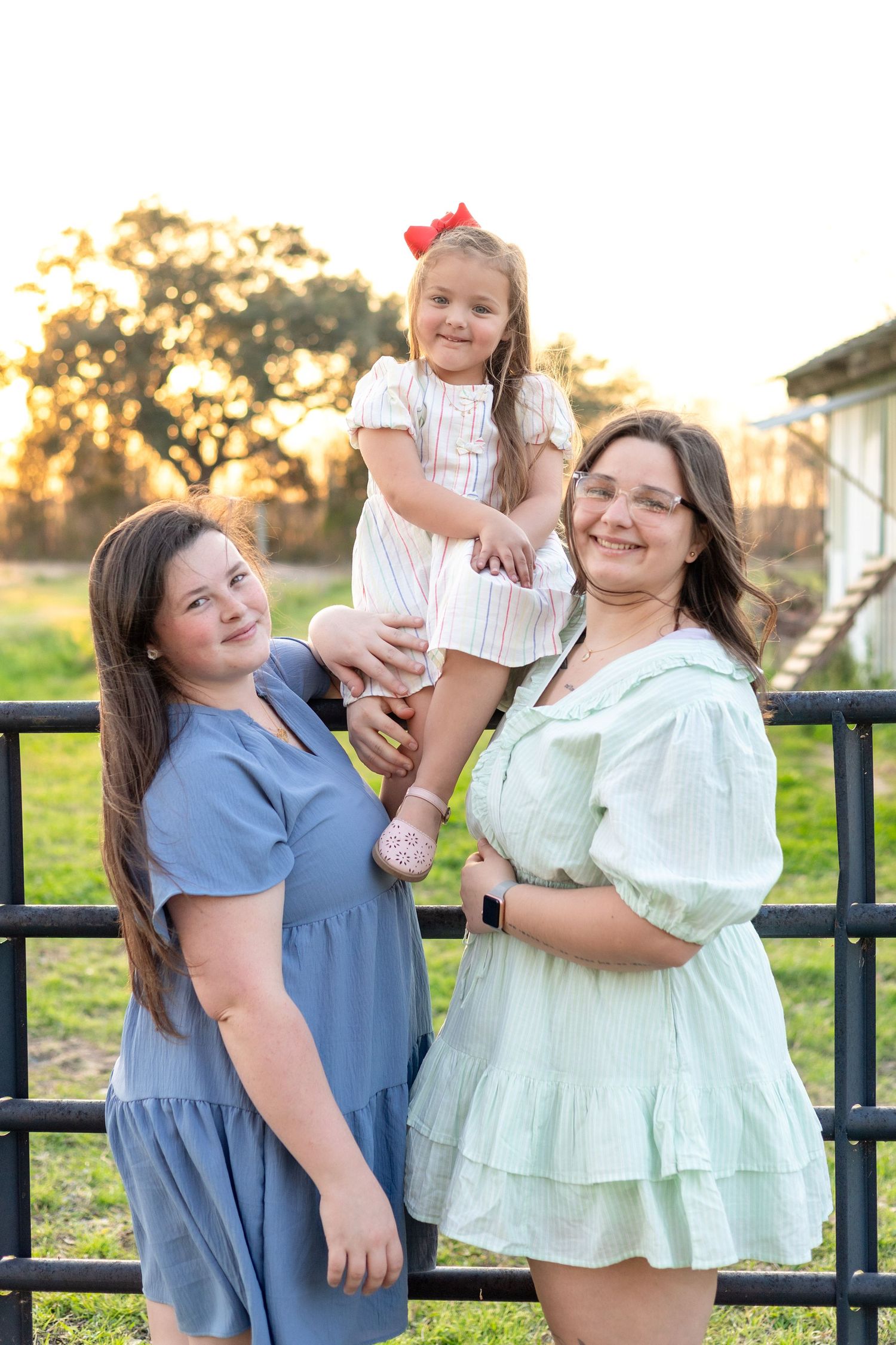 Two women and a young girl in dresses pose by a fence at golden hour on a farm.