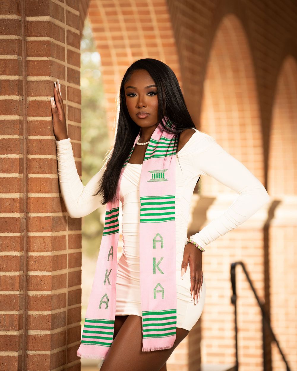 Graduate in white dress with decorative green and pink stole poses against brick wall.