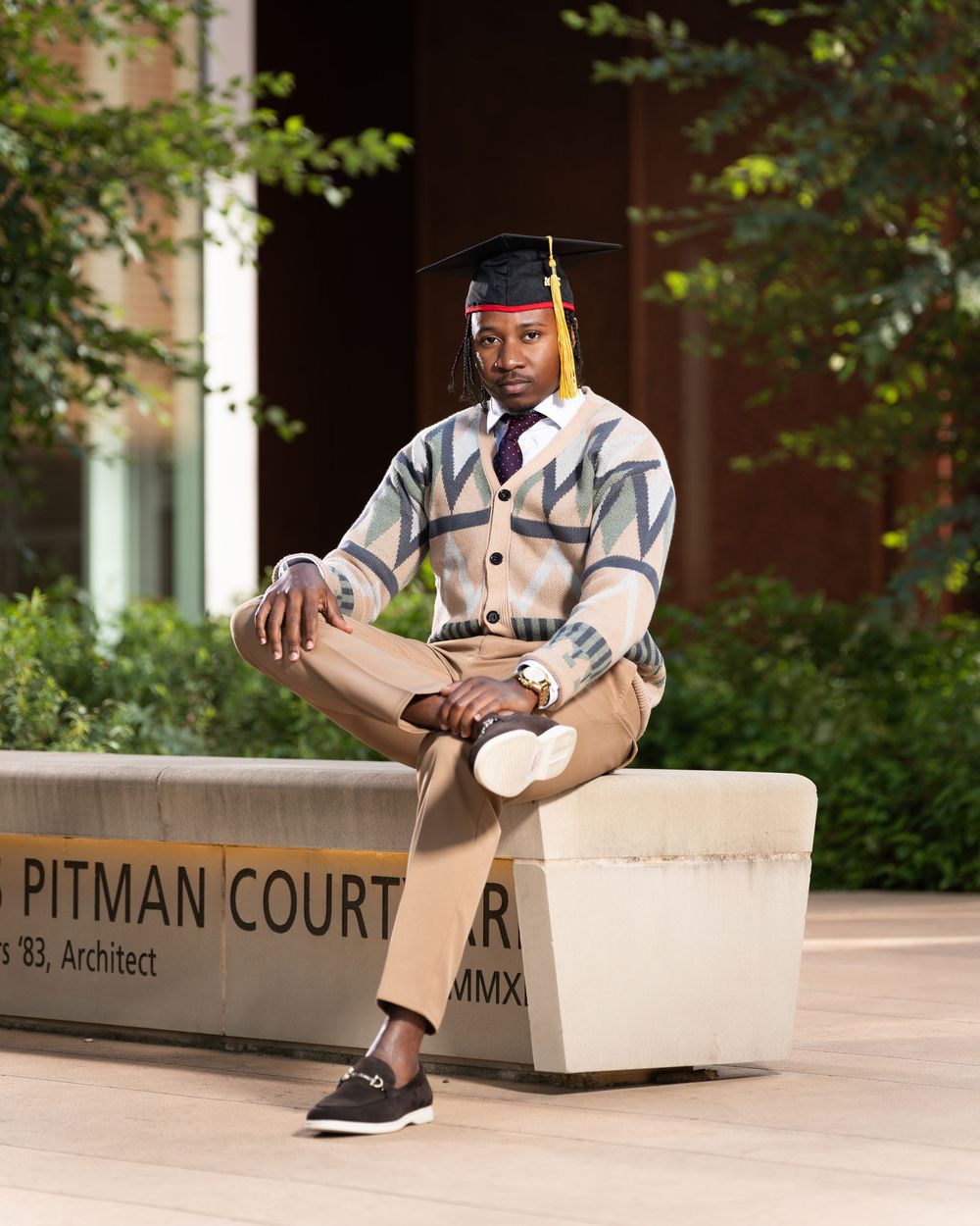 Graduate in academic regalia sits casually on stone bench at Pitman Court.