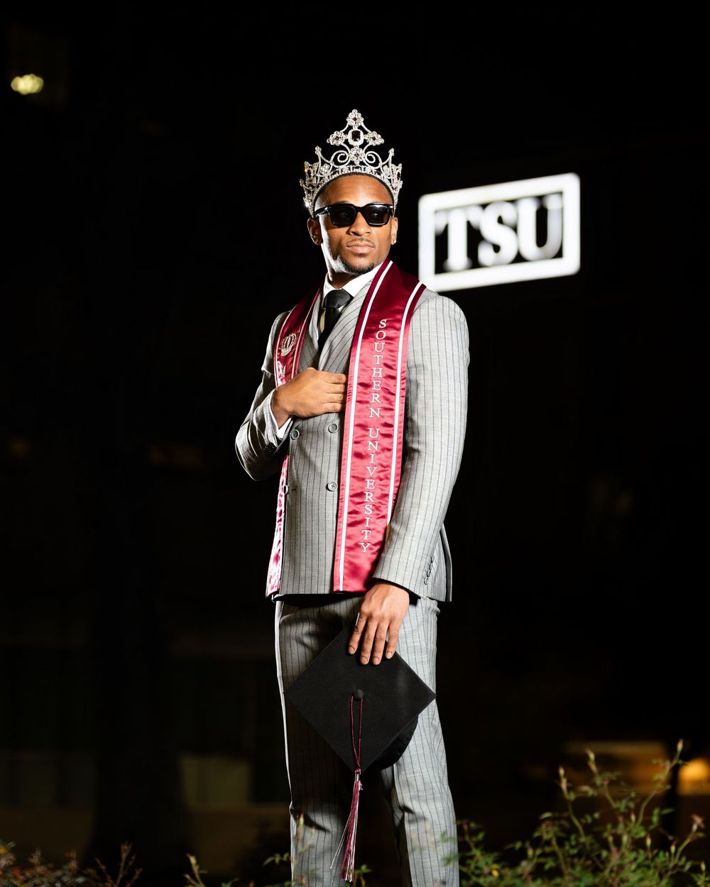 Graduate in pinstripe suit and pink sash poses for nighttime photos.