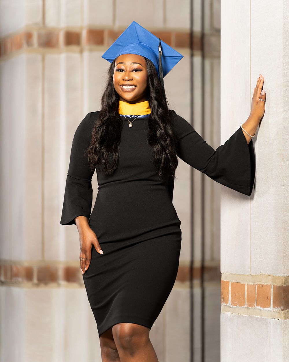 Graduate in black gown and blue cap poses near brick wall during graduation photoshoot.