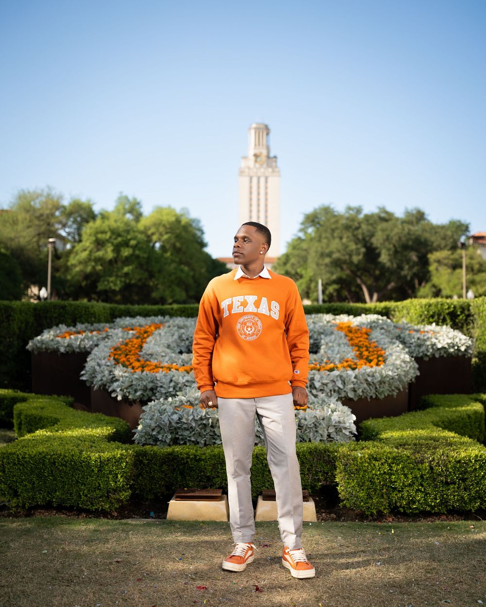 Student in orange Texas sweatshirt standing in front of campus tower with floral landscaping.