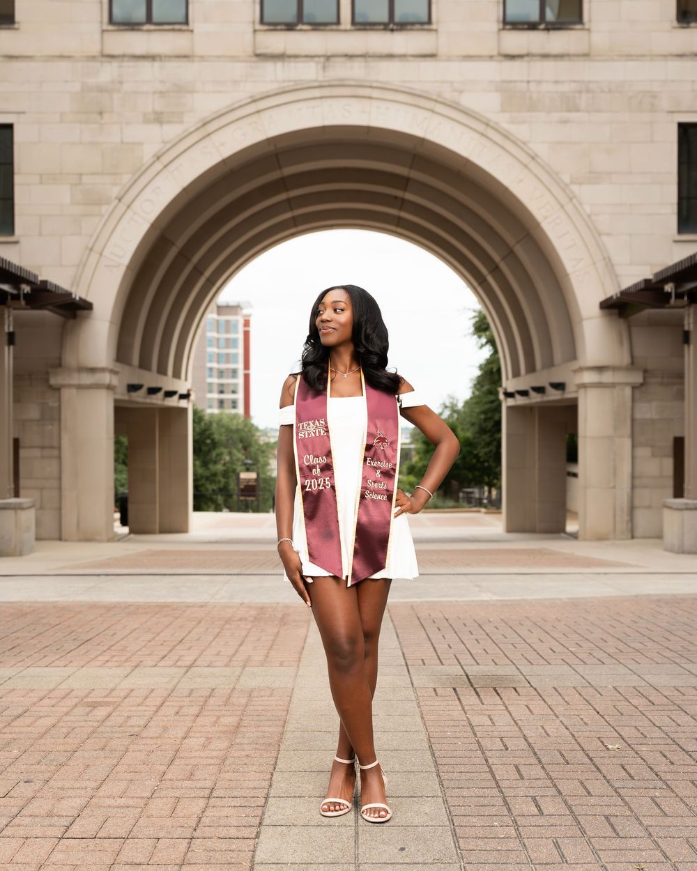 Graduate in white outfit and graduation stole standing under stone archway on campus.