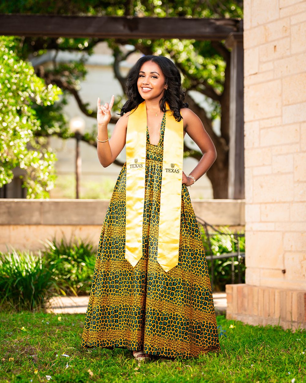 Graduate in vibrant yellow and brown patterned maxi dress poses outdoors with graduation stoles.