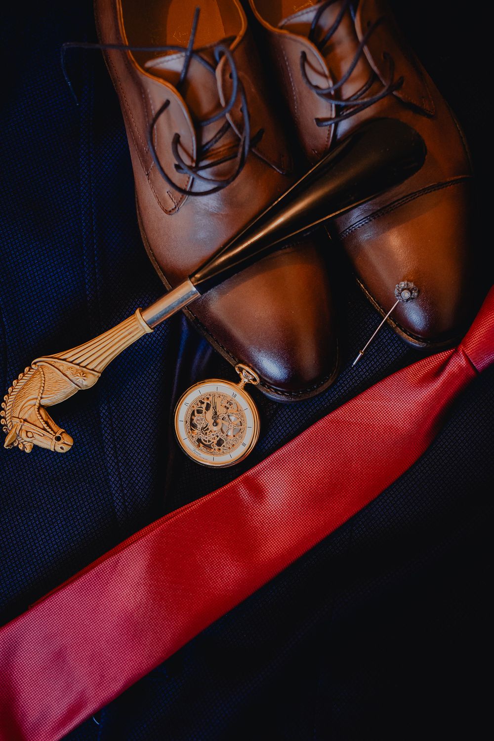 Brown leather dress shoes with a pocket watch and red tie on dark fabric.