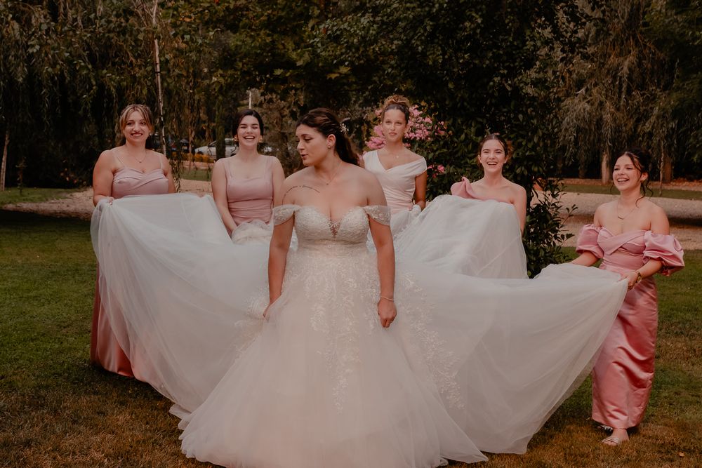Wedding party in pink dresses holding up the bride's white gown outdoors.