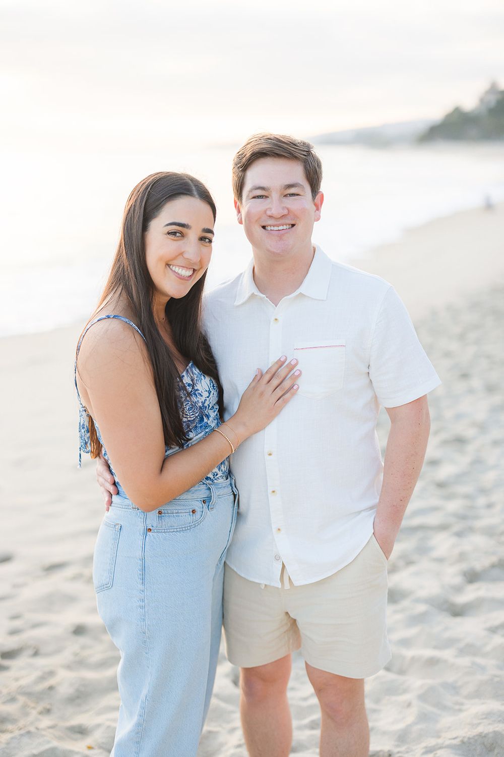 A happy couple embraces and laughs together on a sunny beach wearing light blue and white casual attire.