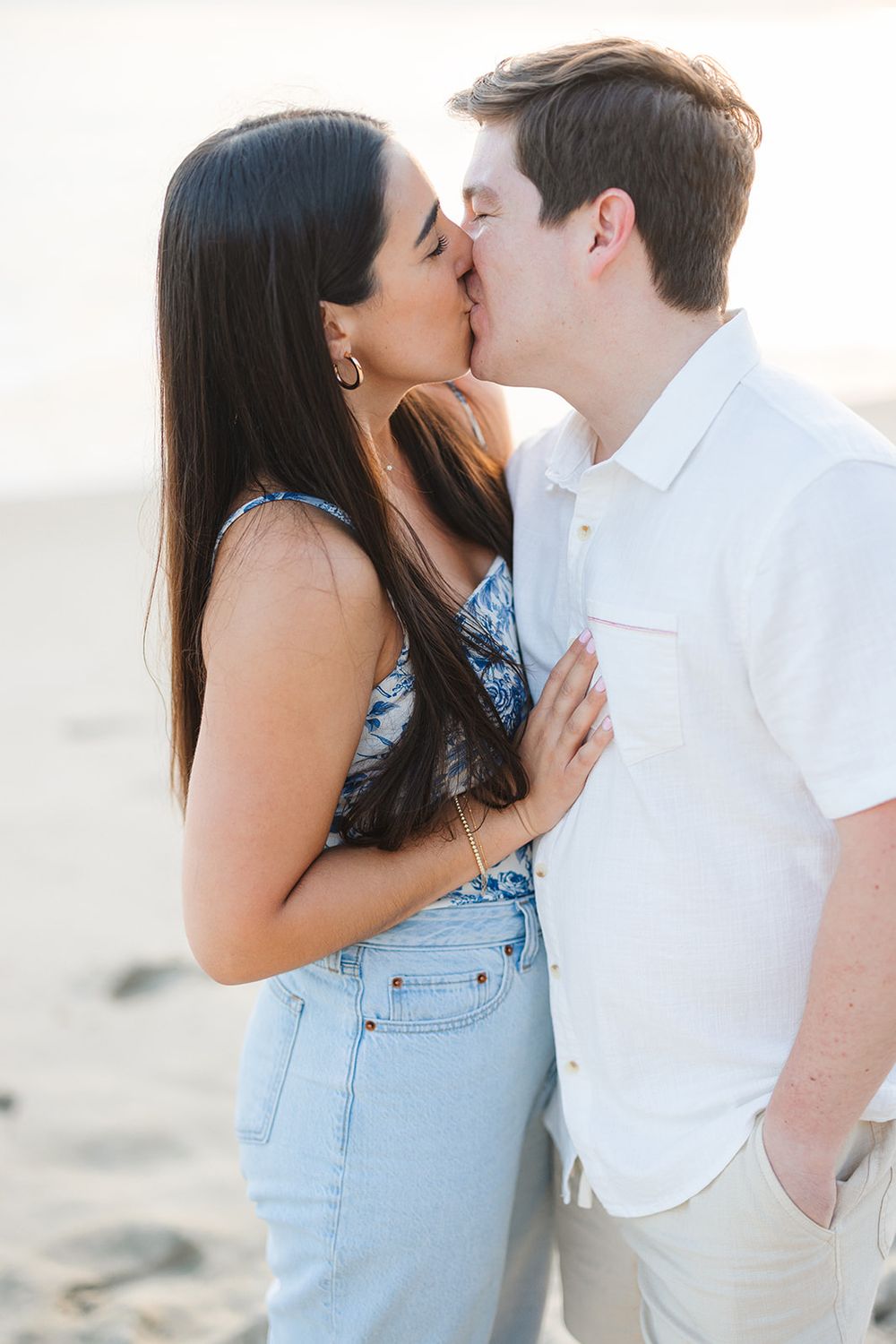 A couple shares a romantic kiss on the beach while dressed in light summer clothing during golden hour lighting.