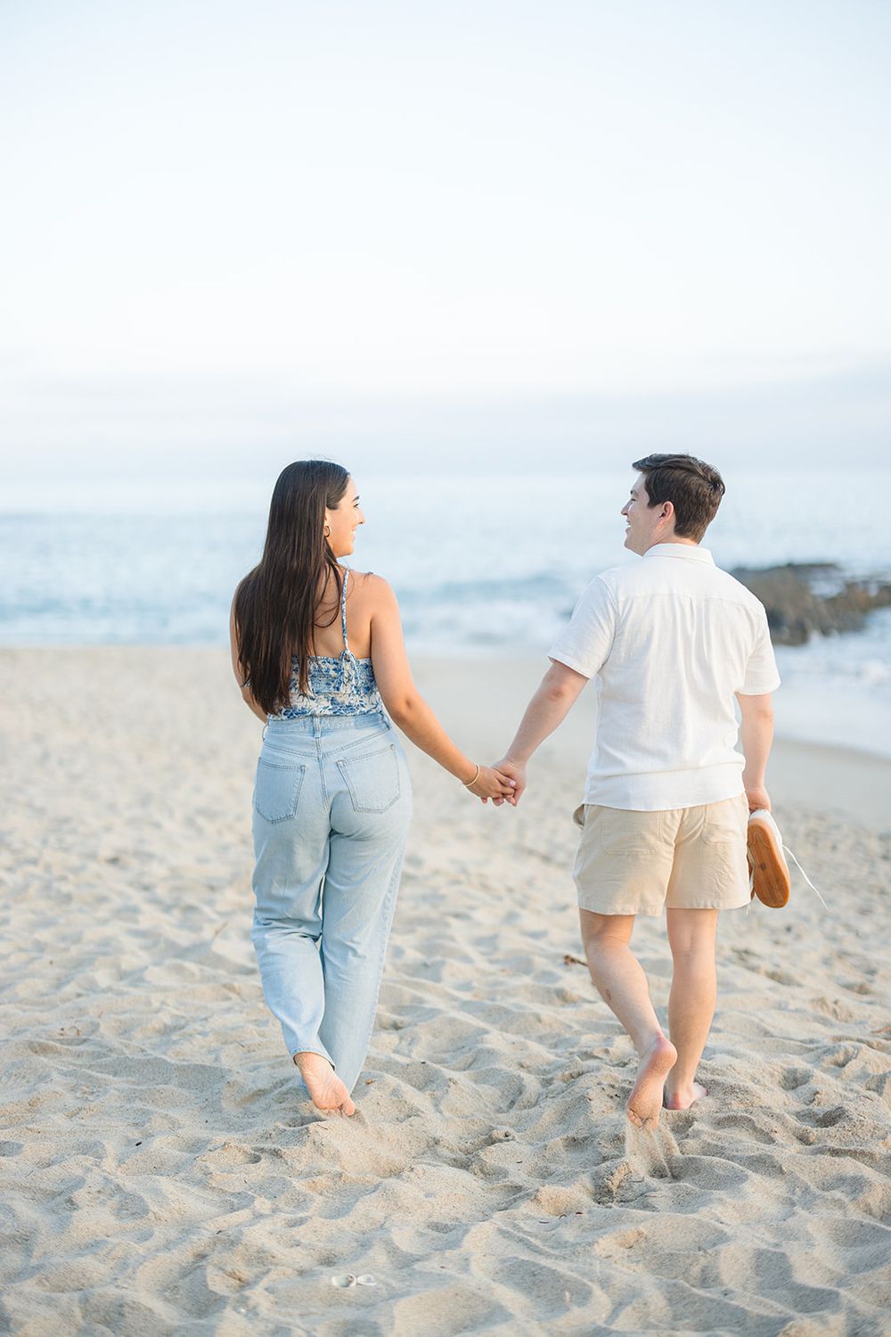 A couple walks barefoot hand in hand along a sandy beach at sunset wearing casual summer attire.