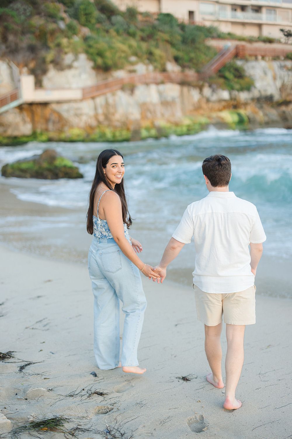 A couple walks barefoot holding hands on a beach with cliffs and turquoise waters in the background.