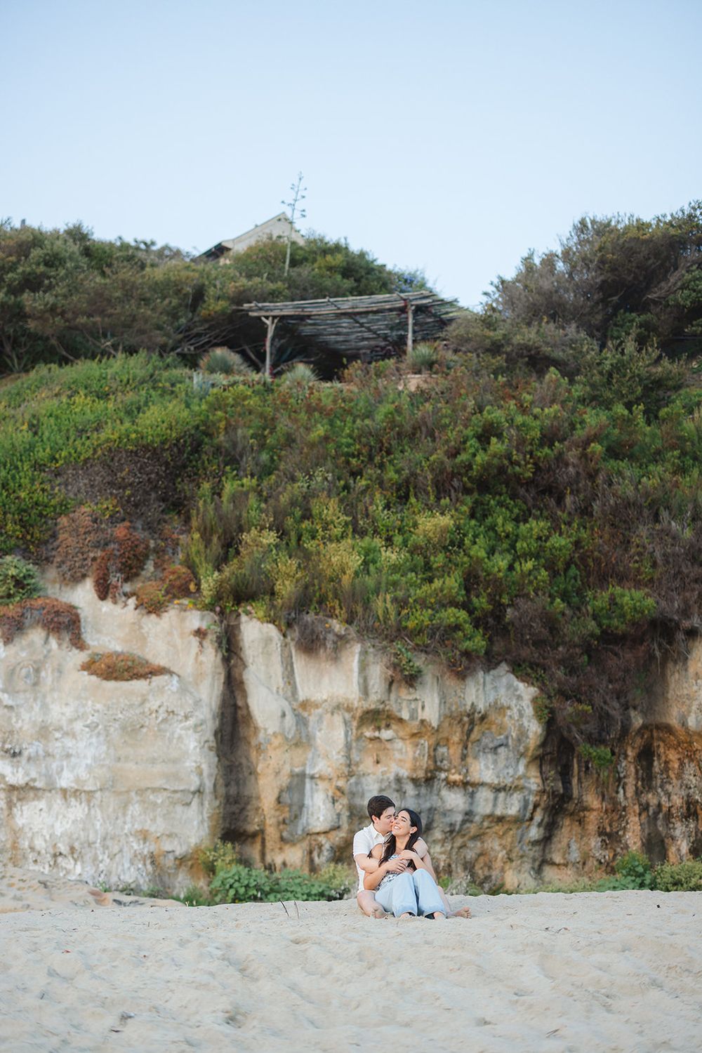 A couple sits together on a sandy beach against dramatic white limestone cliffs and green vegetation.