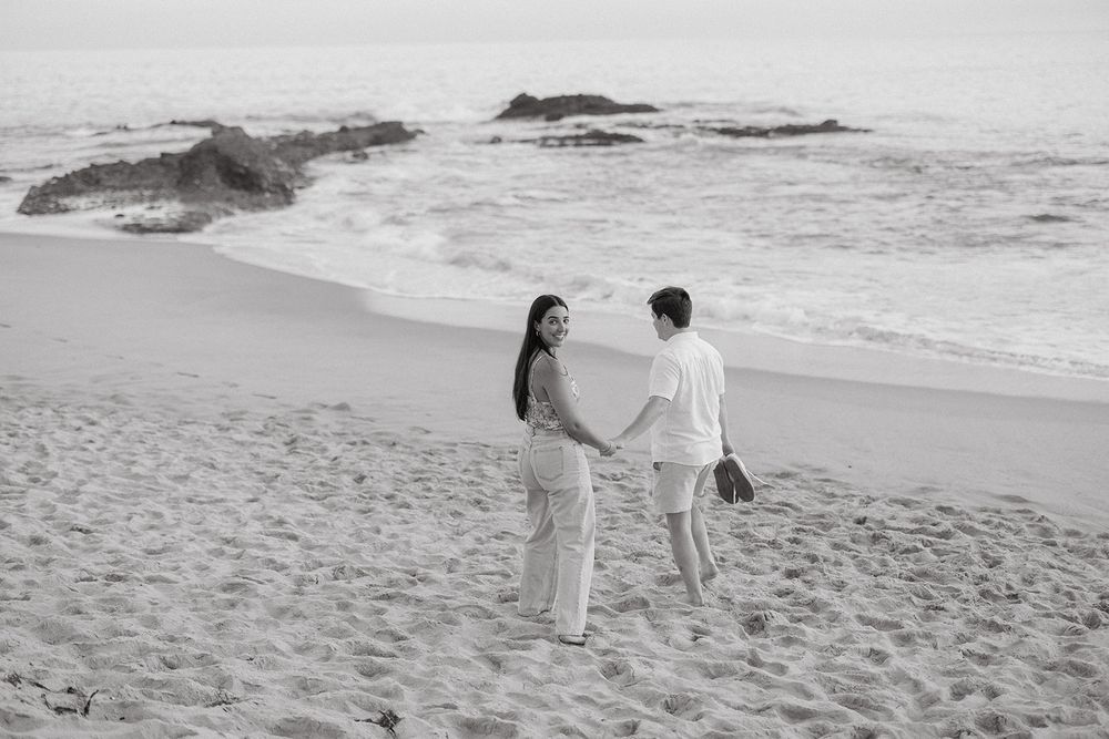 Black and white panoramic image of people walking along a beach shore with waves and rocks in the background.