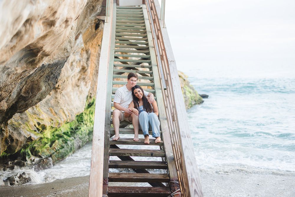 Someone sits on wooden stairs leading down to a rocky beach with waves crashing along the California coastline.