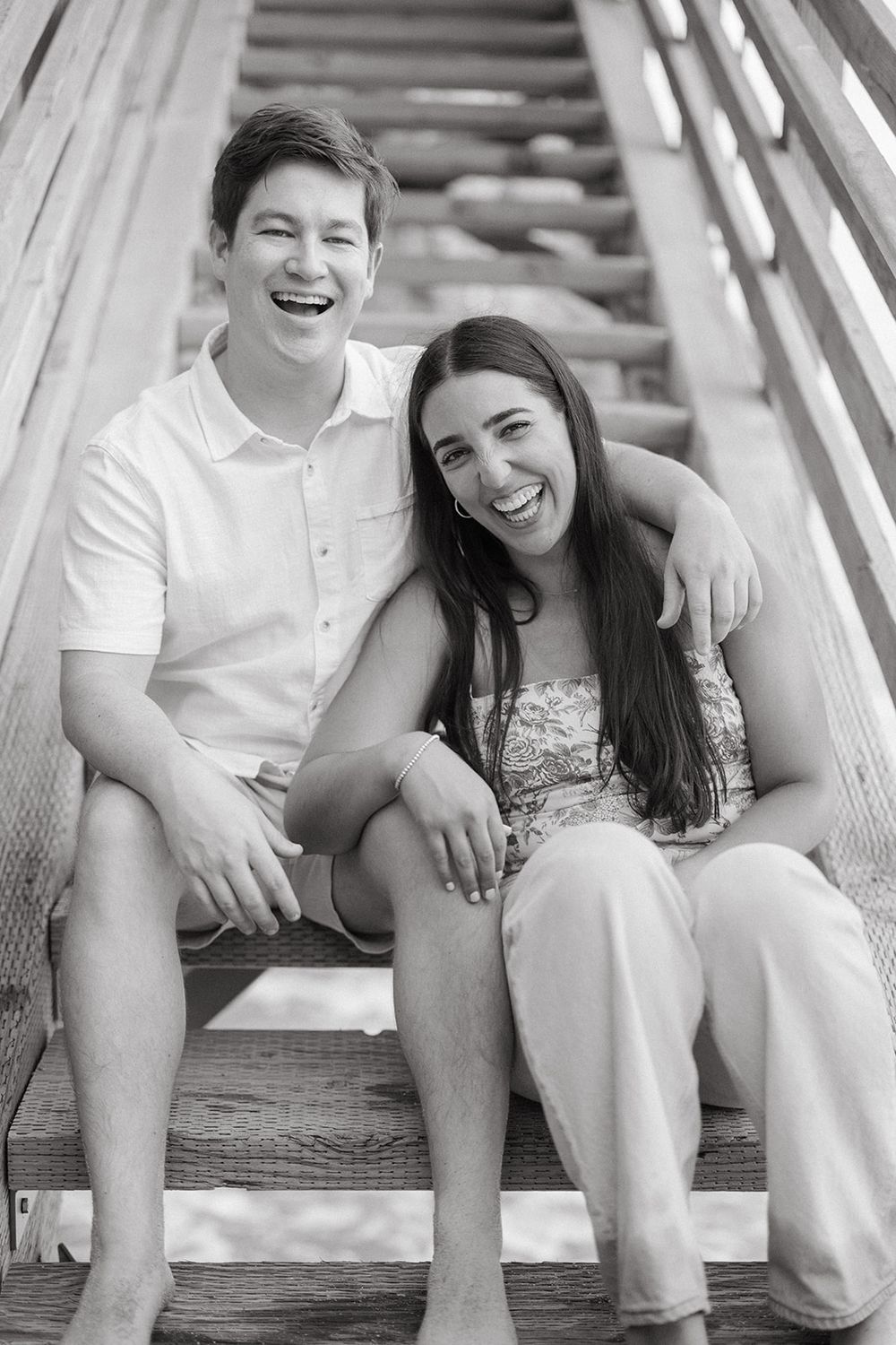 Happy couple in white outfits laughs together while sitting on wooden steps in a candid moment.