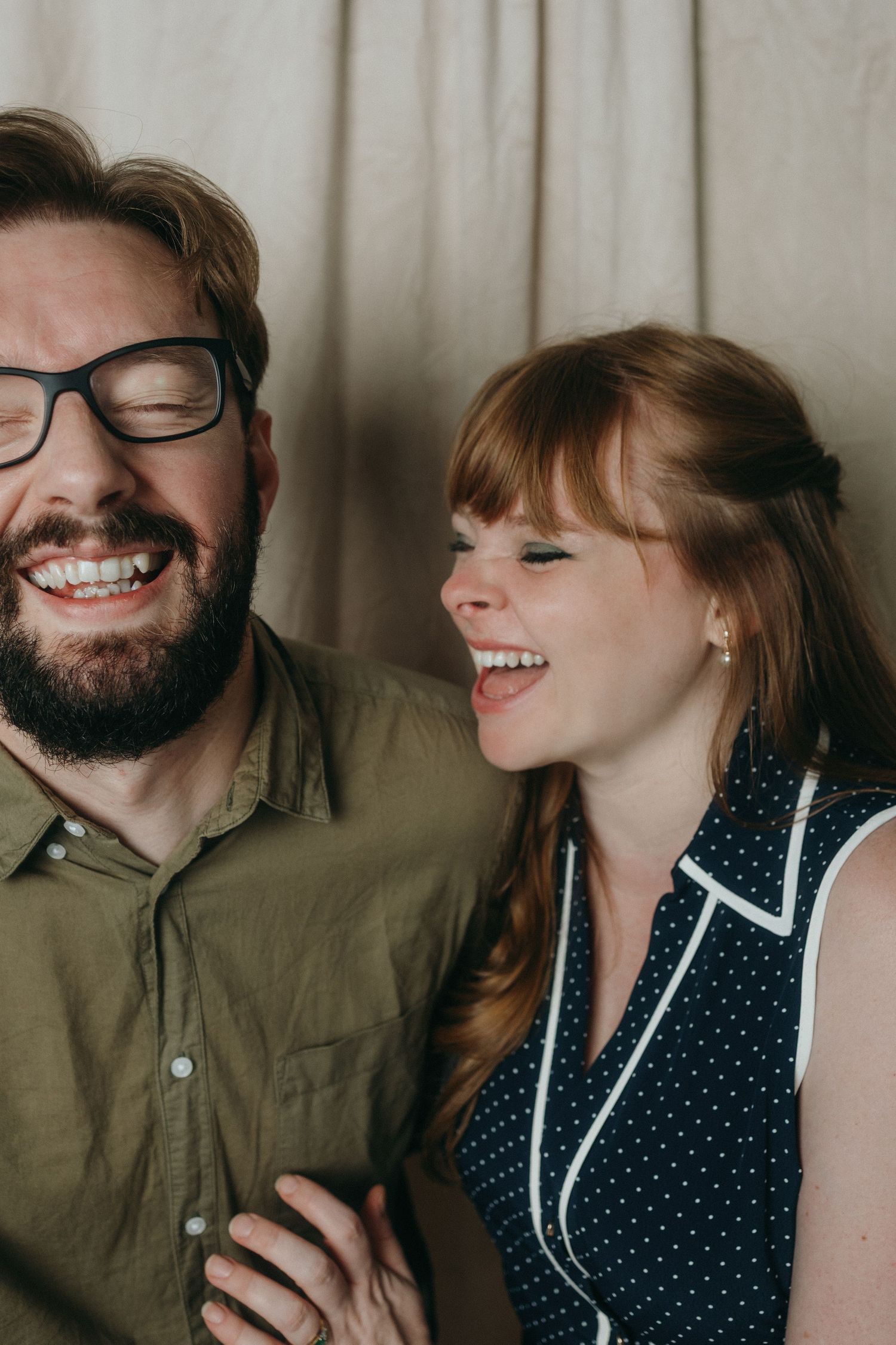A joyful couple shares a laugh together, with him in glasses and olive shirt and her in a polka dot dress.