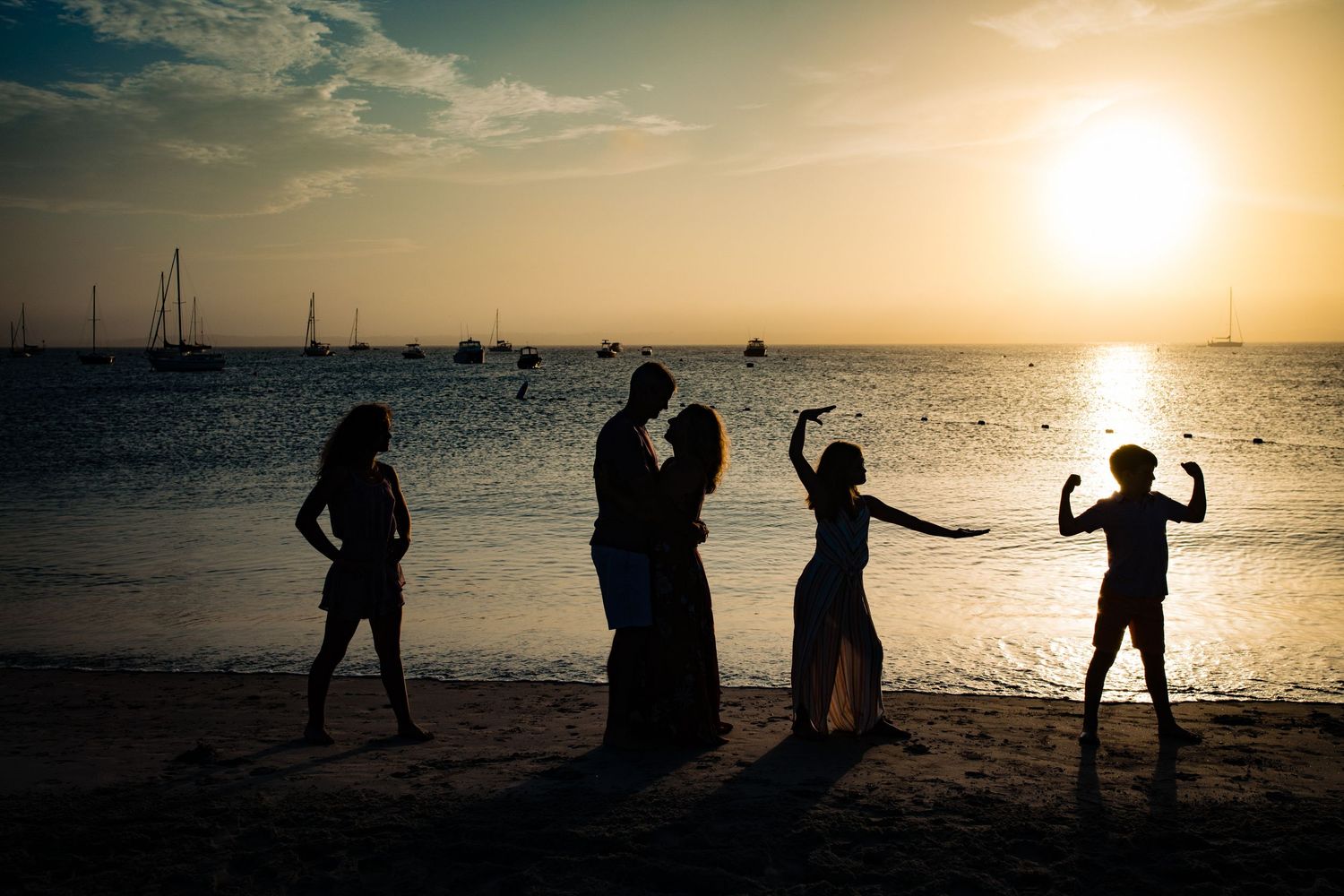 Silhouettes of people dancing and posing on a beach at sunset with boats in the harbor.