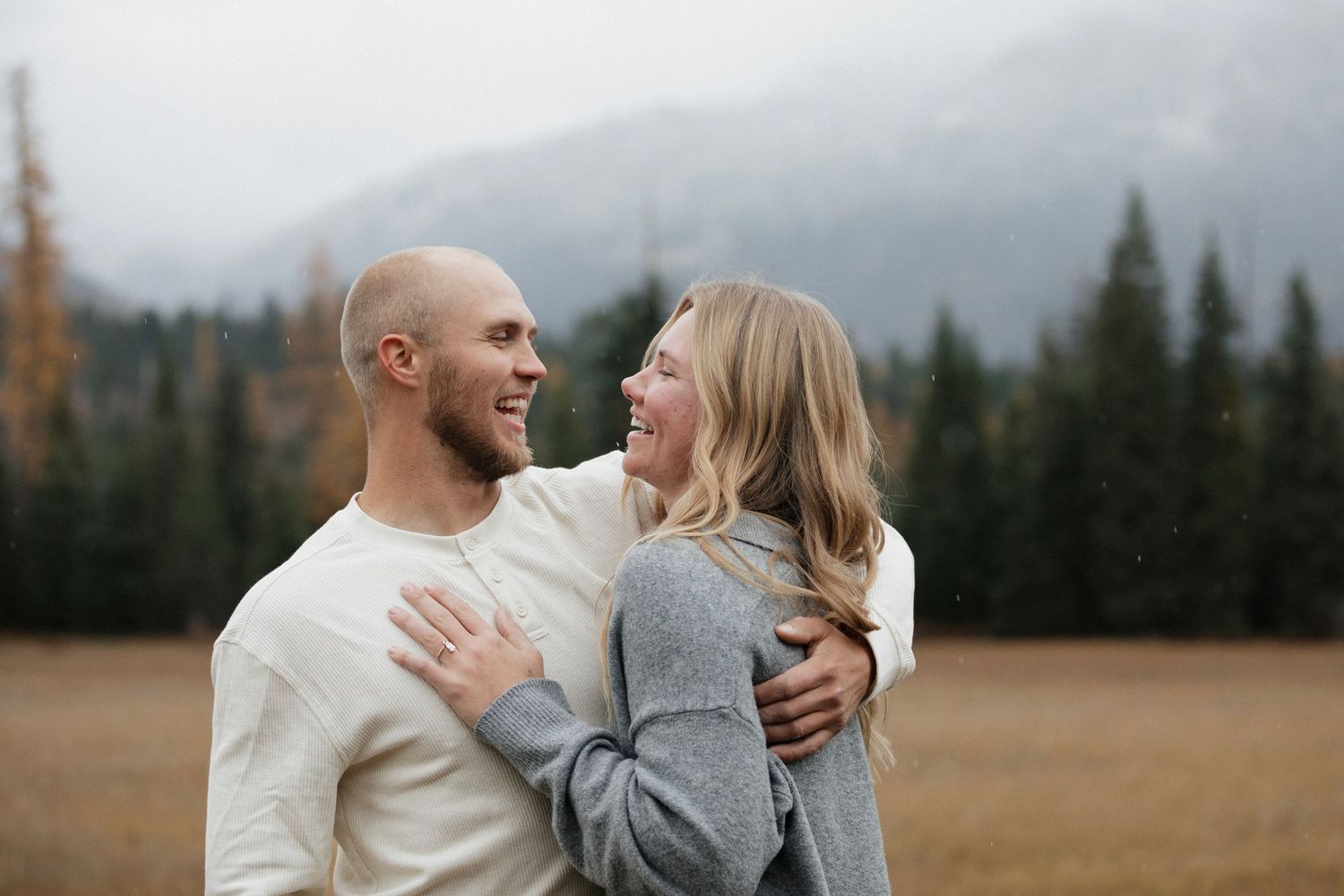 Two people share a joyful moment together while standing in a mountain field at sunset with evergreen trees in background.