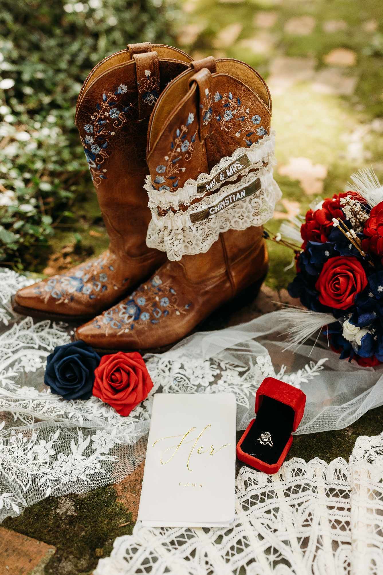 Brown leather cowboy boots with lace veil and red roses next to engagement ring box on rustic surface.