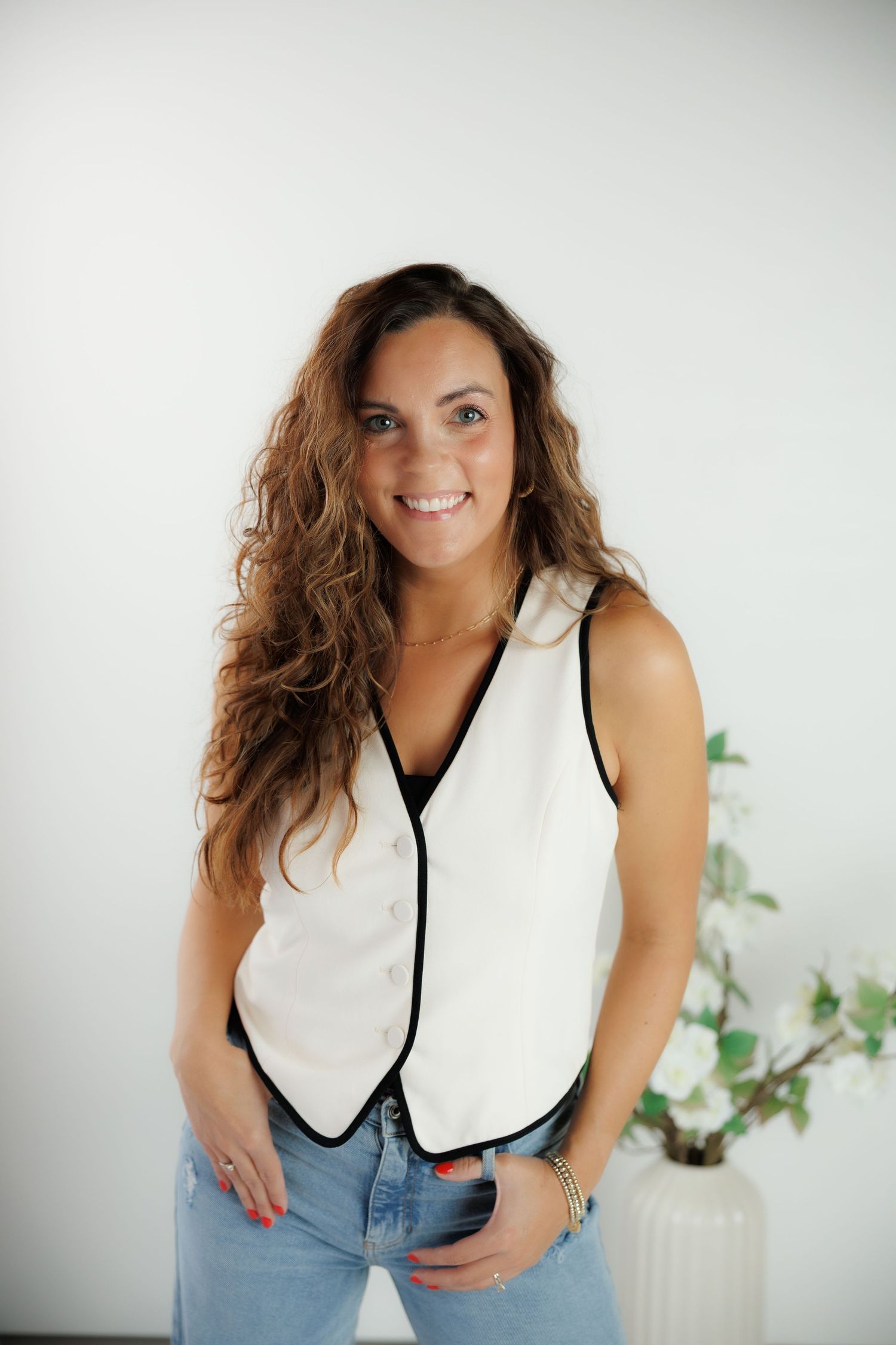 Professional studio portrait of someone wearing a white sleeveless top with dark trim and light denim jeans against a bright wall.