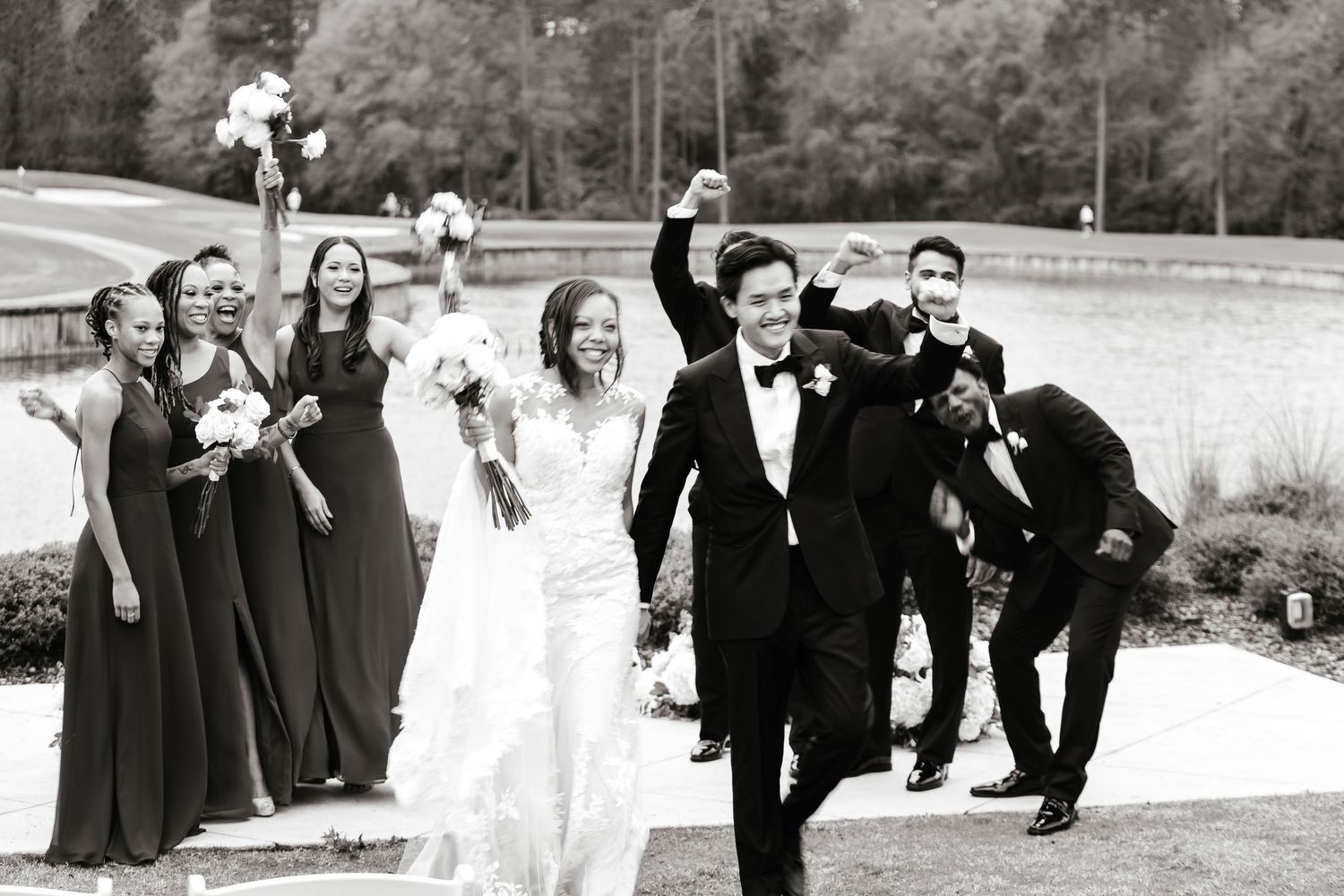 A wedding party walks along a path outdoors, with bridesmaids in dark gowns and groomsmen in formal attire in black and white.