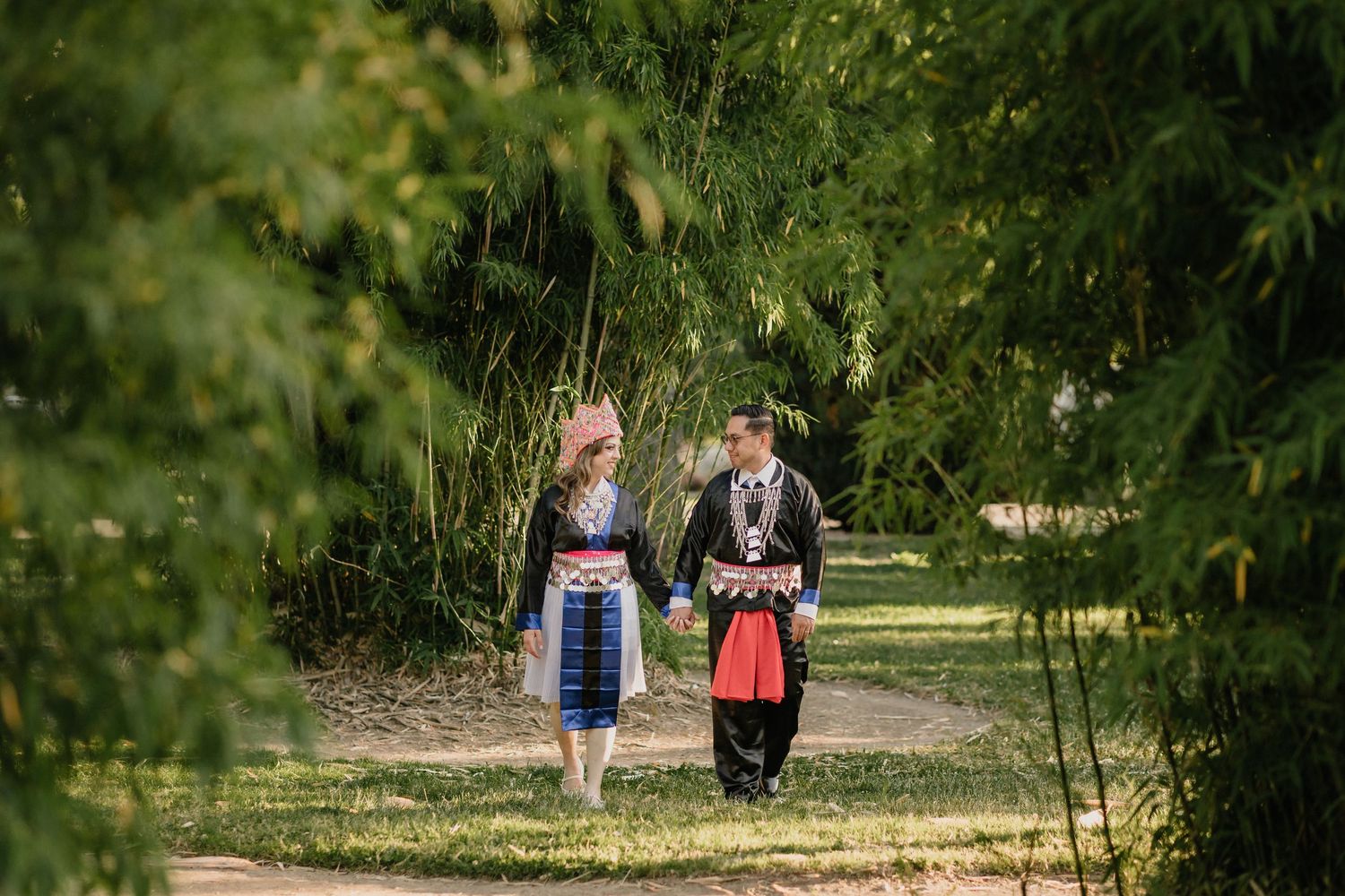 Couple walk together along a tree-lined path in a green park on a sunny day wearing traditional clothes