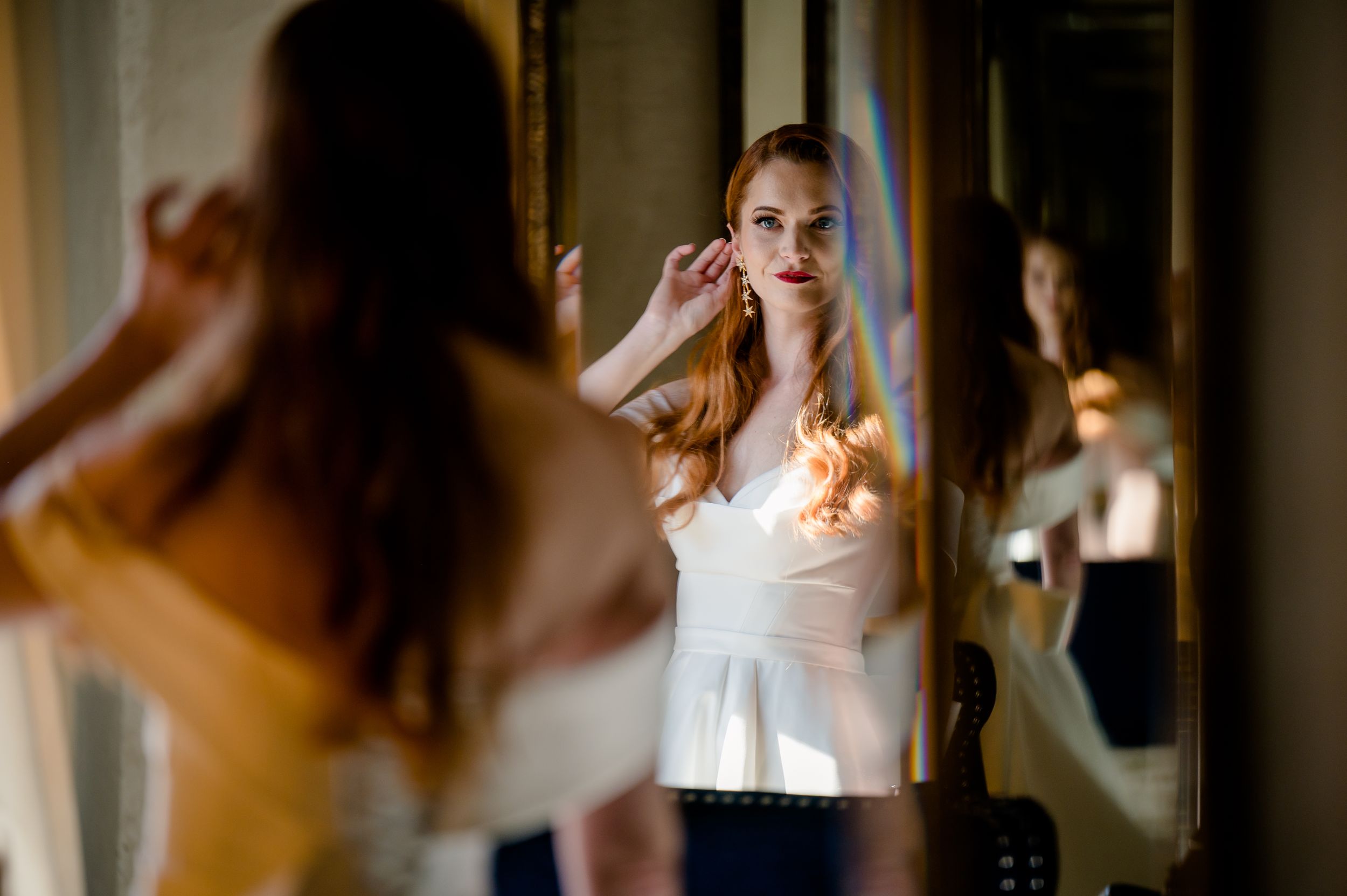 Person putting on earrings while getting ready in front of mirror with warm lighting and reflections.