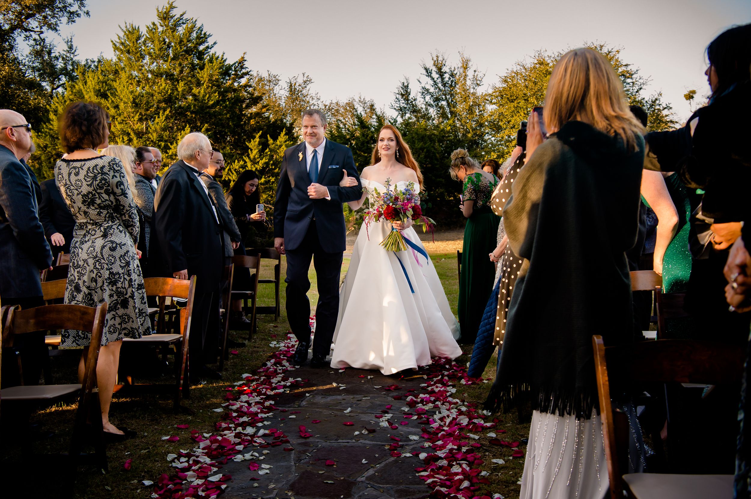 Wedding attendees stand alongside a flower petal-lined aisle during an outdoor autumn ceremony.