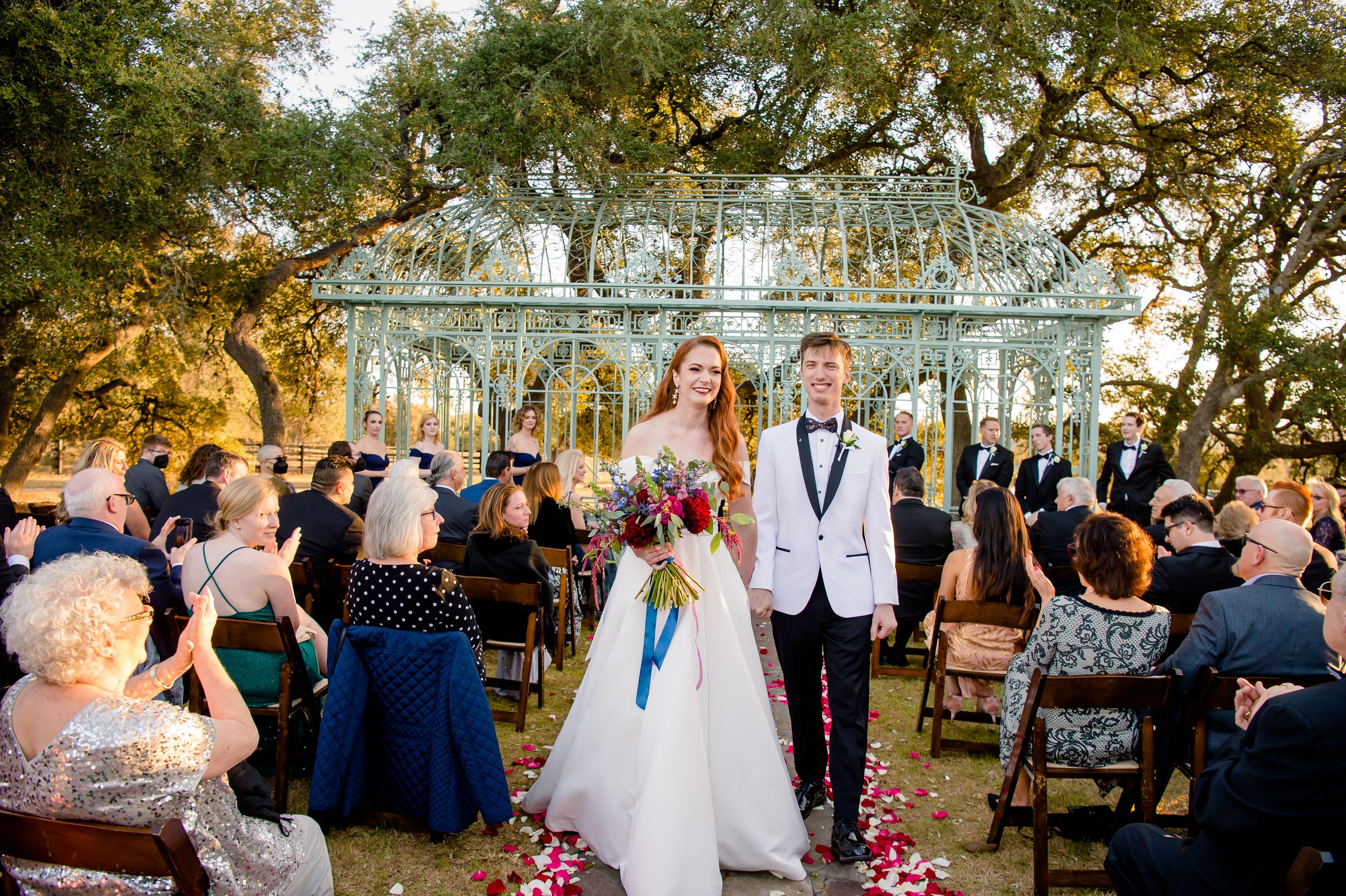 Wedding celebration with guests throwing confetti and rose petals at an outdoor garden venue during a golden sunset.