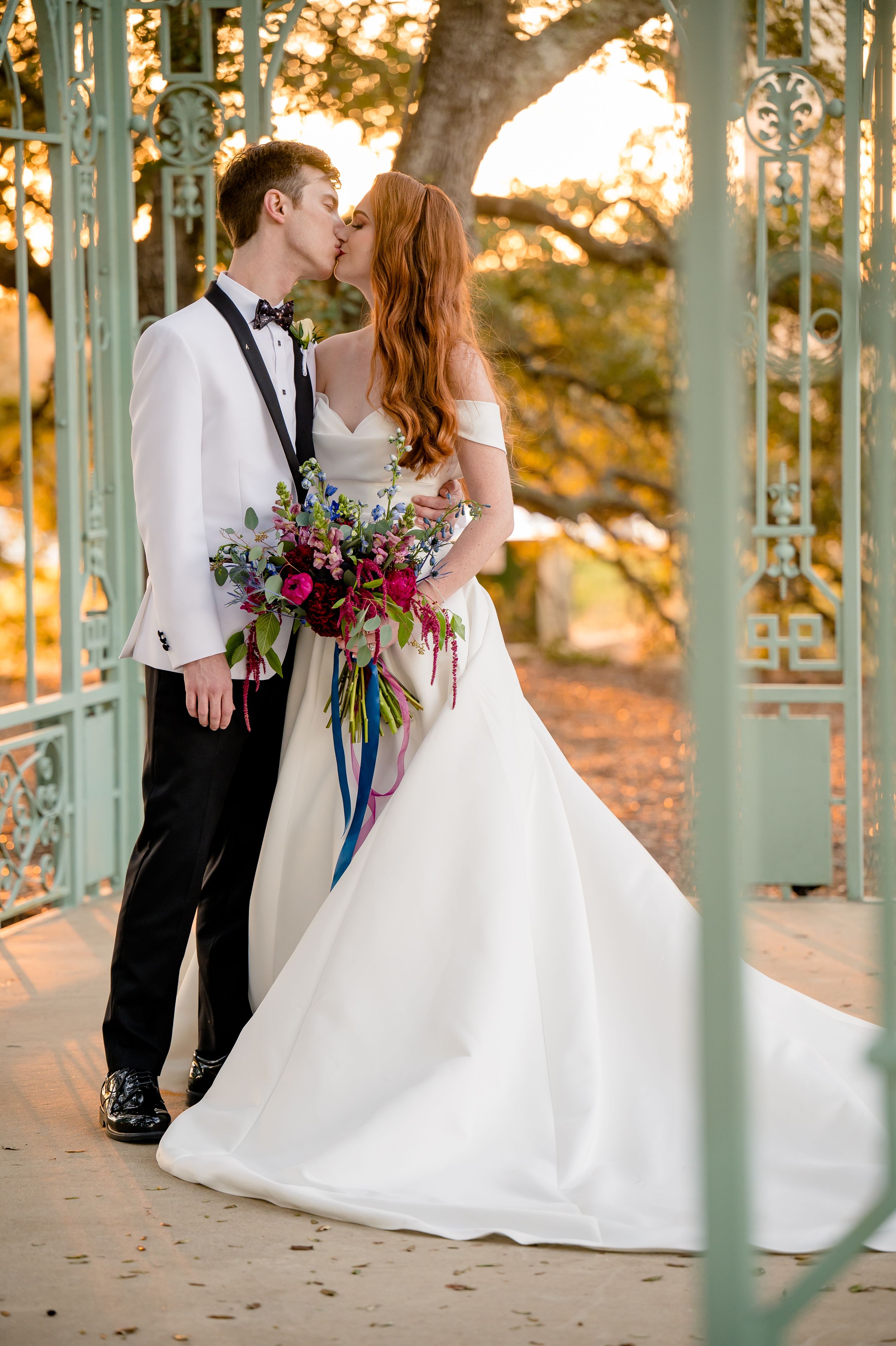 A romantic wedding couple shares a sunset kiss in a teal gazebo while holding a vibrant bouquet with purple flowers.