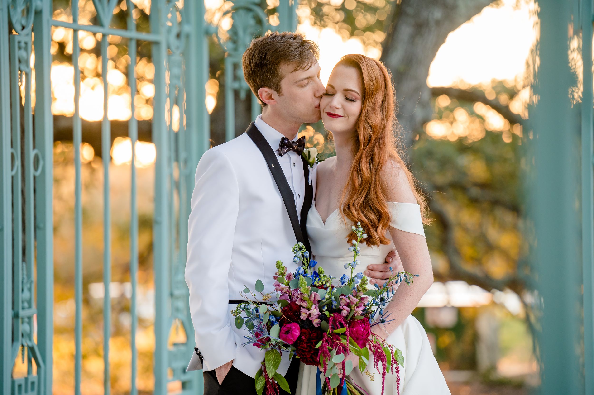 A couple shares an intimate moment at sunset in a garden gazebo with the bride holding a vibrant burgundy bouquet.