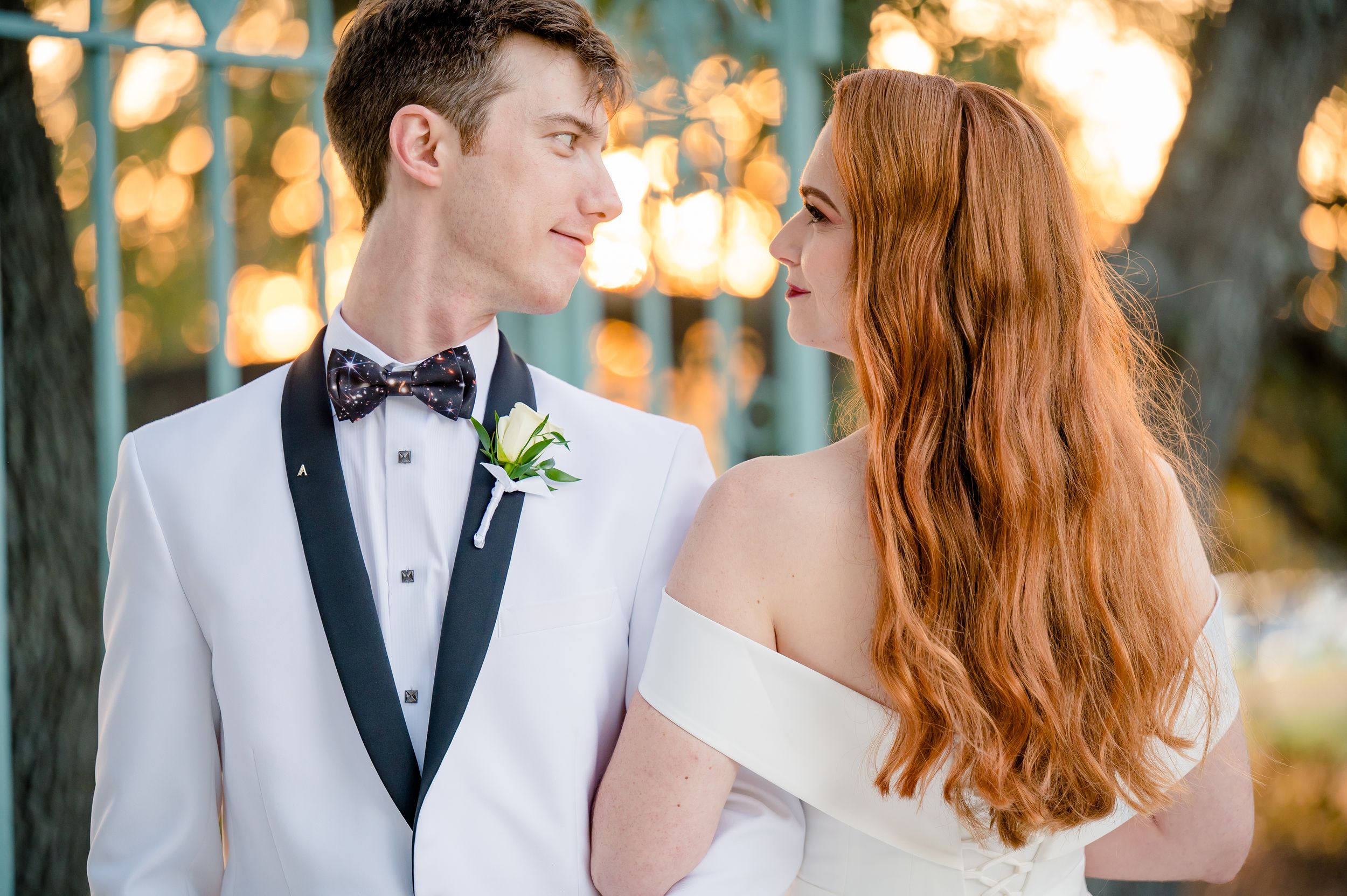 A romantic outdoor wedding photo showing a groom in a white tuxedo and bride with flowing red hair at sunset.