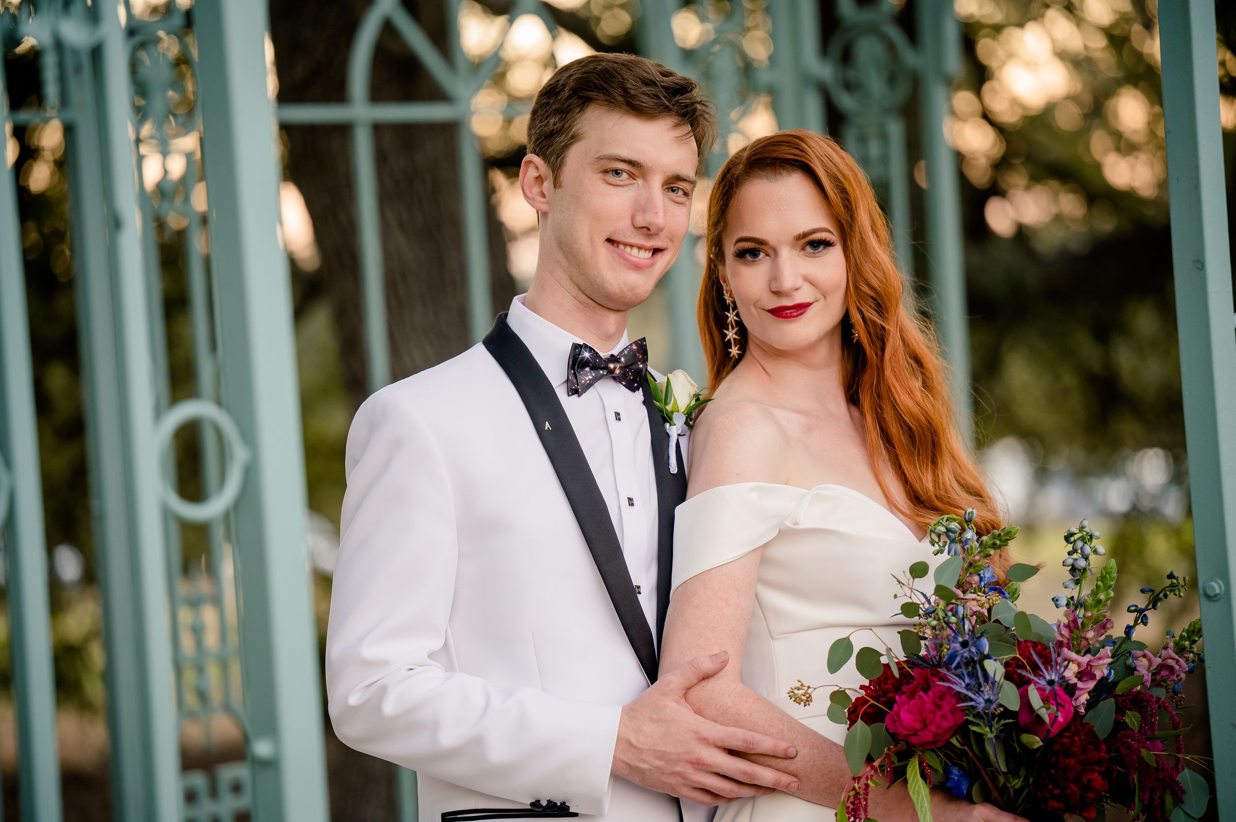 Elegant bride and groom pose together at sunset, with turquoise gazebo and colorful floral bouquet.