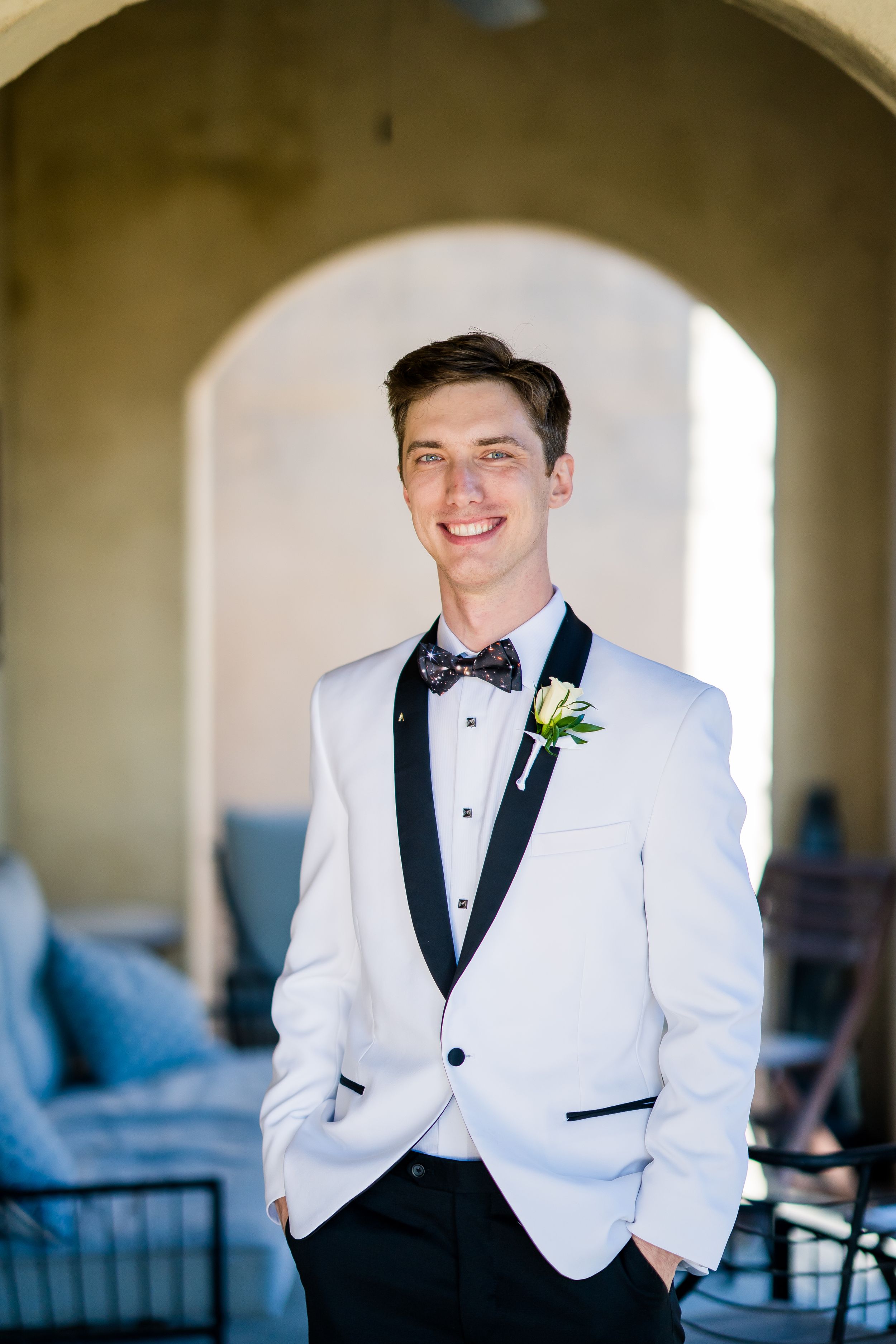 A groom in a white tuxedo jacket with black lapels smiles while posing for formal wedding photos in a bright archway.