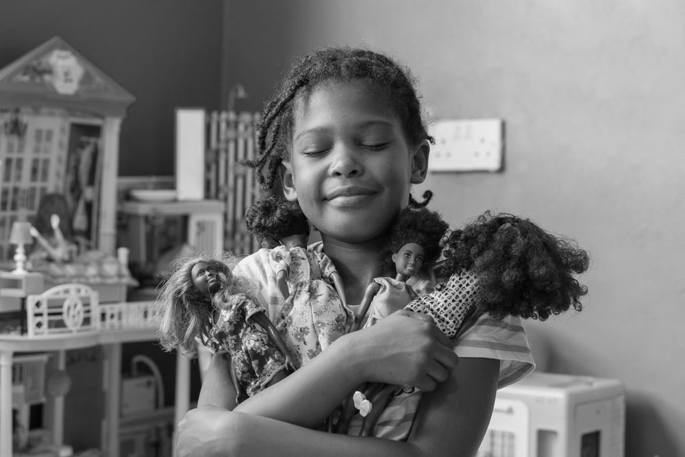 A child smiles while holding a doll near a dollhouse and other toys.