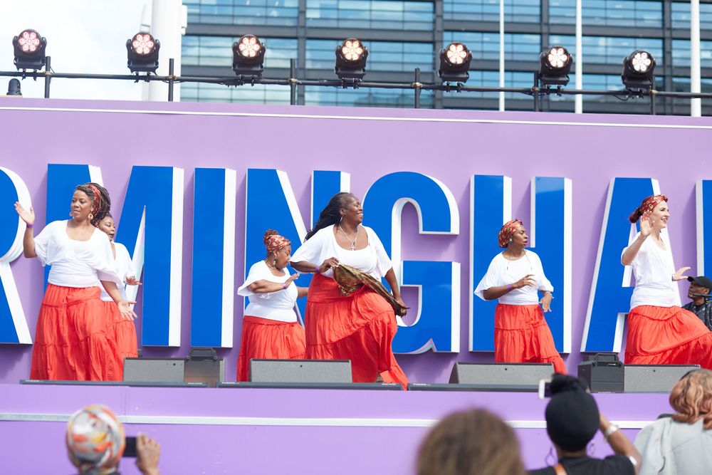 Performers in red skirts dance on stage at an outdoor event in Birmingham.