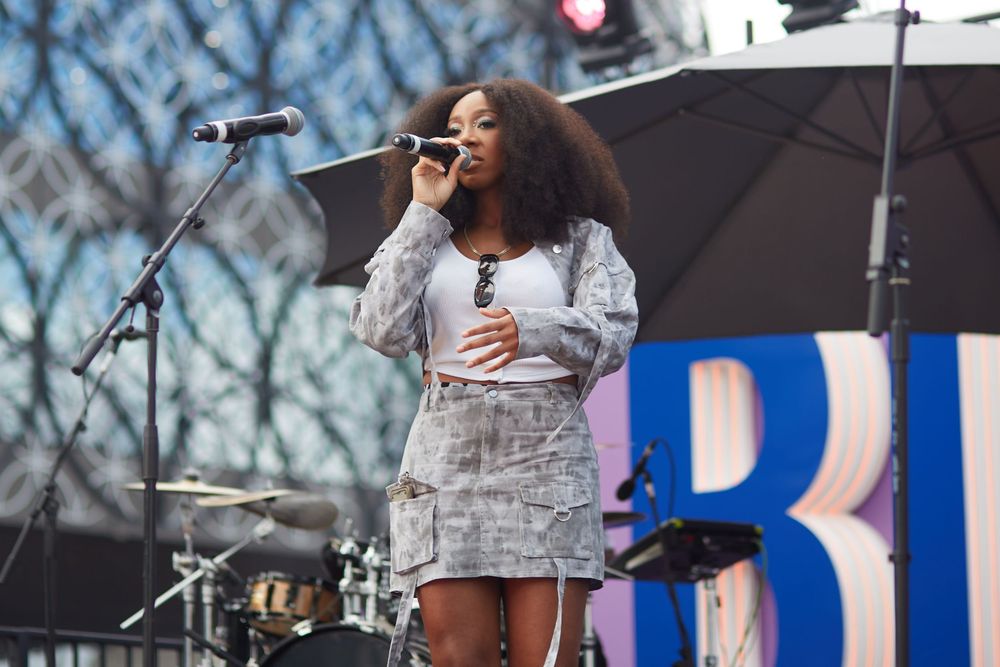 A performer singing on stage at an outdoor festival with trees in the background.